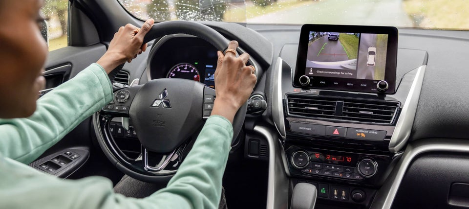 Hands on the steering wheel of a 2023 / 2024 Mitsubishi Eclipse Cross Compact SUV. The screen shows the Multi-View Camera System. 