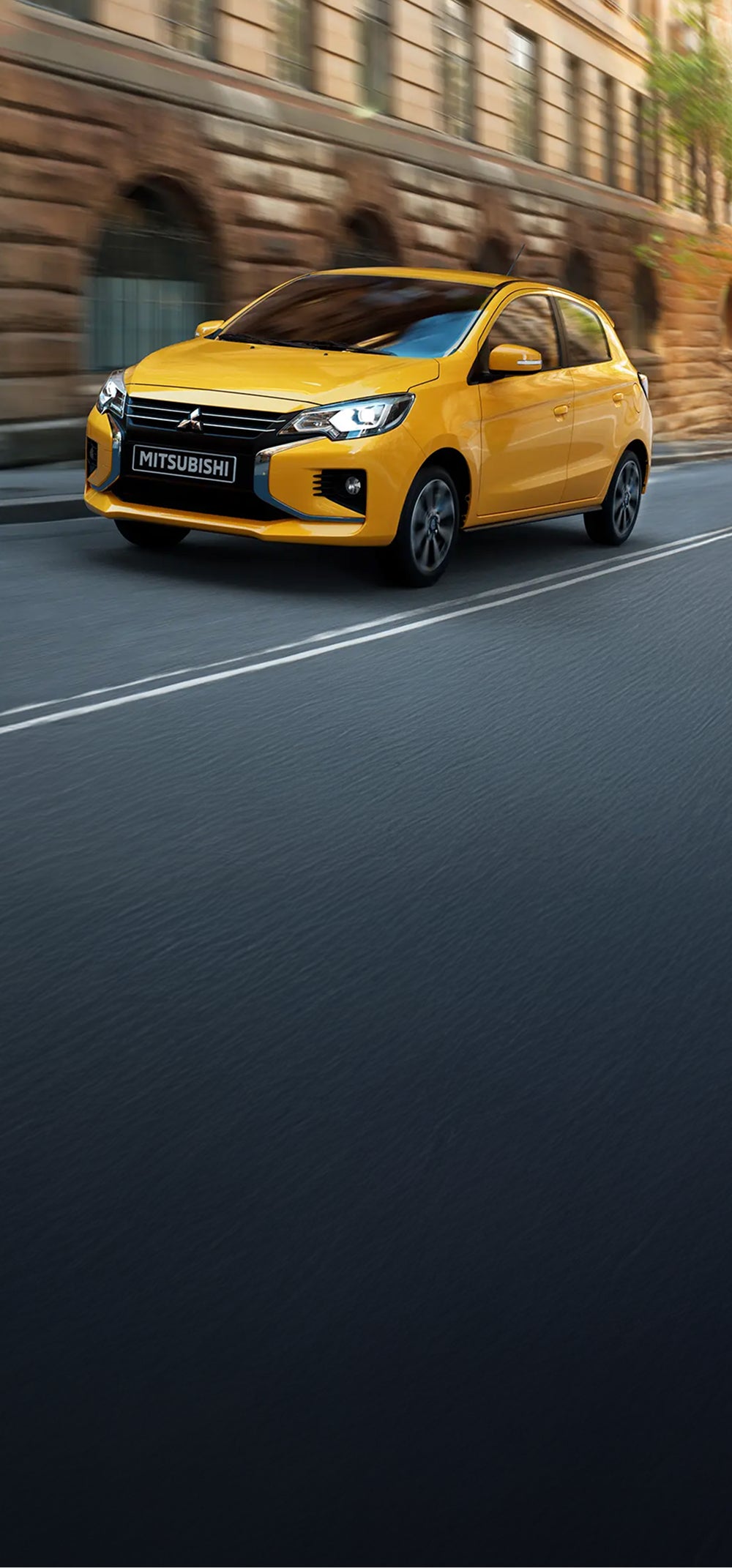 A yellow Mitsubishi Mirage driving on a city street in front of brick buildings.